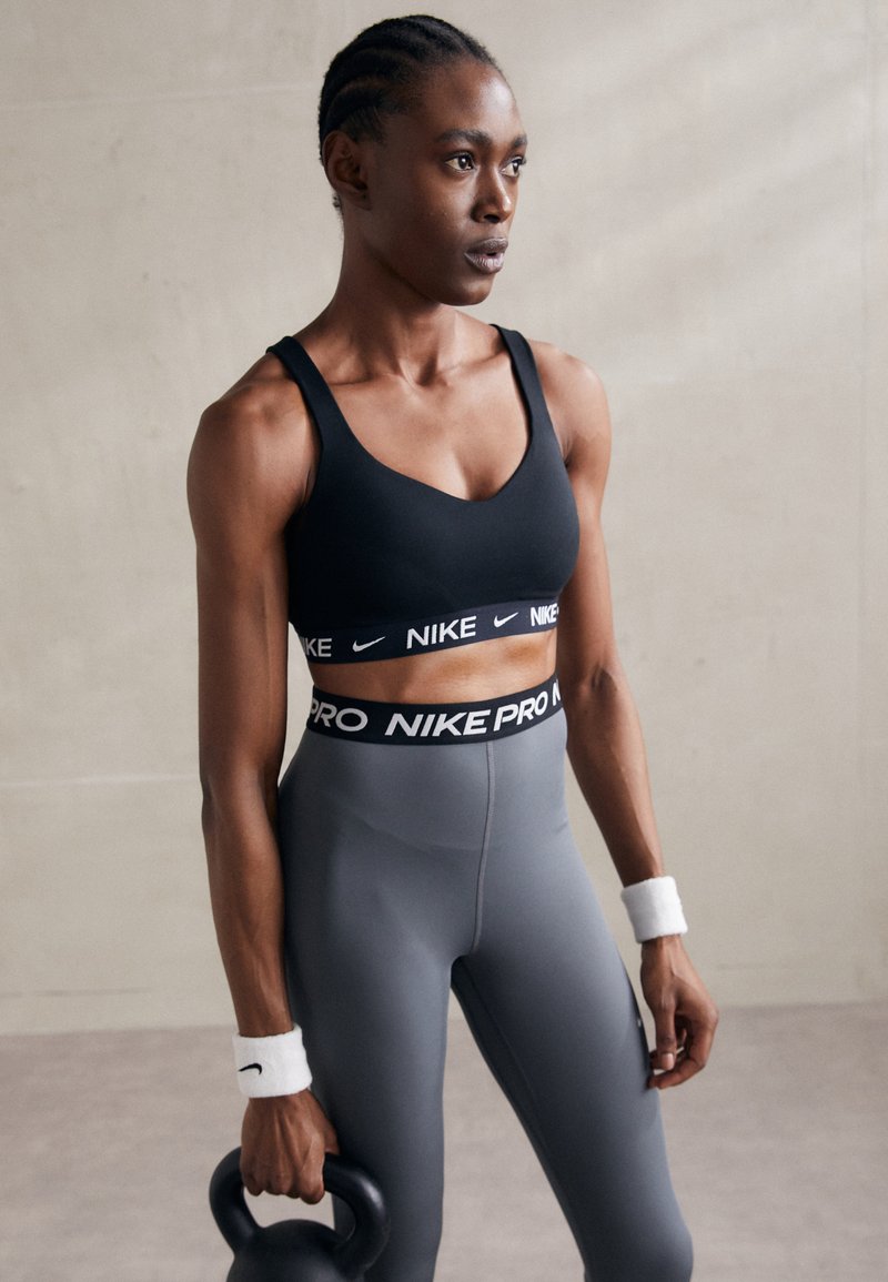 Svart sport-BH og grå tights med bred midje. Materialet ser ut til å være elastisk. Modellen holder en kettlebell, iført hvite håndleddsbånd.