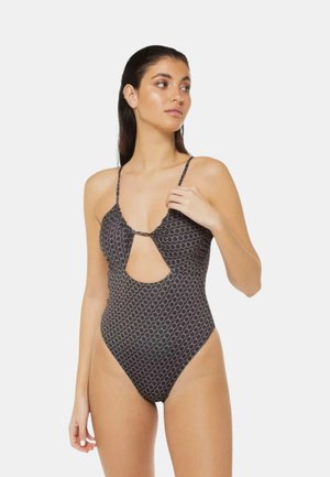 Donna con un costume intero nero con motivo circolare e apertura frontale, mentre sistema la bretella, guardando di lato su uno sfondo neutro.