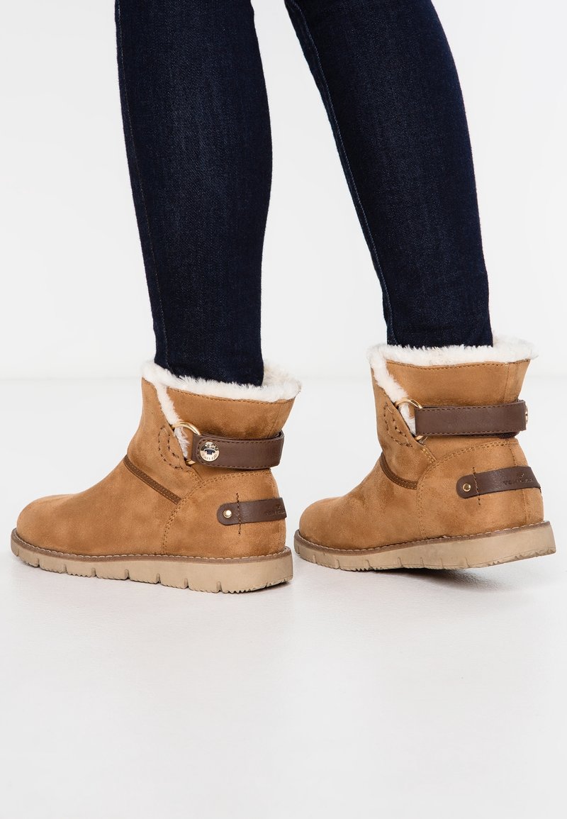 Bottines en daim marron avec une bordure en fourrure blanche douce, dotées d'une lanière en cuir marron et d'une boucle en métal, reposant sur une semelle en caoutchouc texturée.