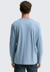 Hellblaues Langarmshirt mit rundem Ausschnitt, Rippung am Kragen und glatter Textur. Getragen mit dunkelblauen Jeans.