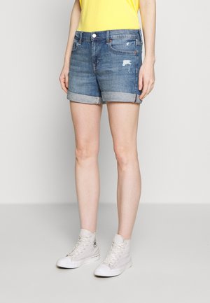 Person iført blå opkørte denimshorts, gul top og hvide høje sneakers, stående mod en ensfarvet baggrund.