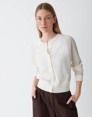 Donna dalla pelle chiara e capelli biondi lisci, indossa un cardigan bianco con bottoni e pantaloni marrone scuro, in piedi con le mani nelle tasche.