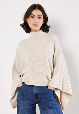 Cremfarbener Strickpullover mit hohem Kragen und gerippter Textur, mit weiten Ärmeln und oversized Passform, kombiniert mit blauen Jeans.