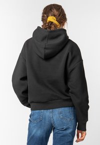 Zwarte hoodie met een comfortabele pasvorm, ribgebreide manchetten en zoom. Gecombineerd met blauwe spijkerbroek. Zicht van achter.