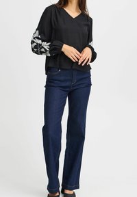 Blouse noire à manches longues ornée de broderies blanches de feuilles sur les manches, associée à un jean droit bleu foncé. Fini simple et texturé.