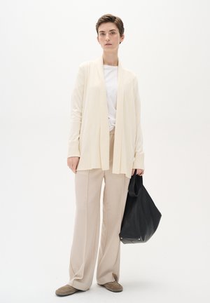 Cardigan lejer de culoare crem cu manșete cu nervuri, purtat cu pantaloni largi bej și o geantă tote neagră. Material neted cu o croială confortabilă.