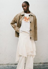 Beige corduroy jas met zwarte knopen, gedragen over een witte graphic tee met oranje ontwerpen, gecombineerd met een ivoren rok en wijde broek.