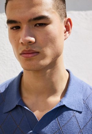 Junger Mann mit kurzen Haaren, der ein blaues Hemd mit Kragen trägt und mit neutralem Ausdruck leicht nach unten schaut, vor hellem Hintergrund.