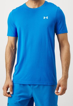 Mężczyzna w niebieskiej krótkiej sportowej koszulce Under Armour i dopasowanych spodenkach na jednolitym tle.