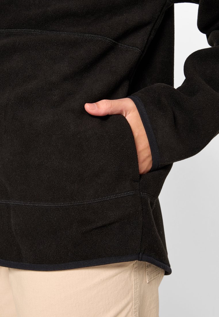 Una mano appoggiata nella tasca laterale di una giacca in pile nera indossata sopra pantaloni beige chiari.