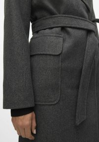 Cappotto di lana grigio scuro con vita regolabile, dettagli di ampie tasche e polsini a coste, caratterizzato da una texture morbida e un design sartoriale.