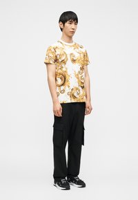 Άνδρας που στέκεται φορώντας λευκό και χρυσό πουκάμισο με κοντά μανίκια και σχέδιο, μαύρο παντελόνι cargo και μαύρα αθλητικά παπούτσια σε απλό φόντο.