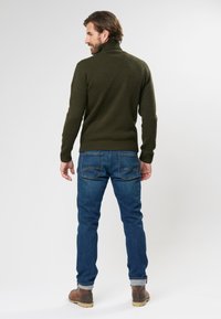Pulover tricotat de culoare verde închis cu guler înalt, asortat cu blugi din denim albastru. Blugii au buzunare la spate cu un design subtil. Botinele maro finalizează aspectul.
