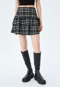Jupe mini à carreaux noir et blanc avec des couches volantes, dotée d'une taille ajustée et d'une silhouette évasée courte, portée avec de grandes bottes noires.