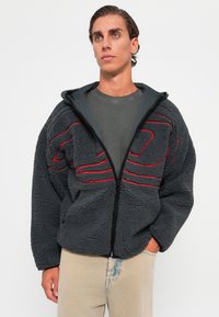 Gray fleece jacket with red stitching accents, zip front, and hood. Features a textured surface and relaxed fit. Worn over a gray t-shirt.