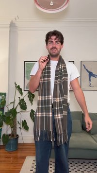 Vlnený šál s hnedo-sivým kockovaným vzorom a bielymi a oranžovými akcentmi. Šál má třásne na koncoch a je prehodený okolo krku.