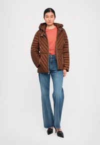 Kvinne iført en brun quiltet kappejakke over en rustfarget skjorte, blå vide benjeans og svarte spisse hæler, stående.