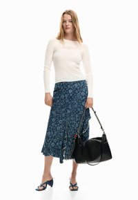 Noor naine, kes kannab valget soonikkoes kampsunit, sinist lillelist midi-seelikut, siniseid kontsakingi ja hoiab suurt musta käekotti ahelatega rihmadega.