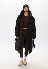 Cappotto lungo e nero imbottito con cappuccio, caratterizzato da sezioni gonfie e una cerniera. Indossato sopra un top corto marrone e leggings coordinati, con scarpe da ginnastica.