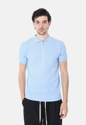 Uomo in piedi contro uno sfondo bianco che indossa una polo a maniche corte in maglia azzurro chiaro con cerniera e pantaloni neri con coulisse bianche.