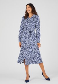 Abito a maniche lunghe e colletto di colore blu navy con motivo floreale bianco. Presenta una vita con cintura e uno spacco laterale. Il tessuto ha una texture liscia.