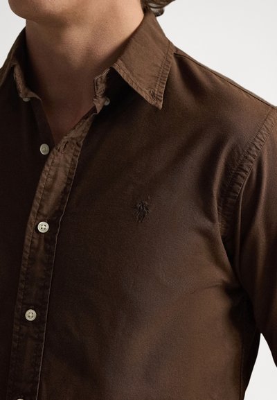 Camisa castanha de botões feita de tecido suave com um brilho subtil. Apresenta uma gola estruturada, botões brancos e um emblema no peito.