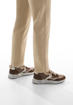 Piernas que llevan pantalones beige y zapatillas marrones con suelas blancas gruesas que tienen grandes detalles de texto gris.