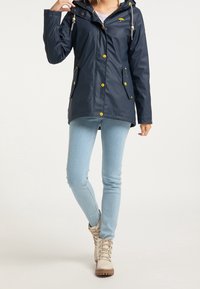 Vattentät marinblå regnjacka med huva, gula knappar och sidofickor. Mjuk textur med kontrasterande dragsnören. Matchad med ljusblå jeans.