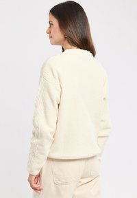 Maglione off-white con texture e motivo a trecce sulle maniche, scollatura rotonda e finitura morbida, abbinato a pantaloni chiari.