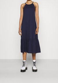 Mörkblå ärmlös klänning med en samlad byst och utflugen flerlagers nederkant, kombinerad med vita snörda kängor.