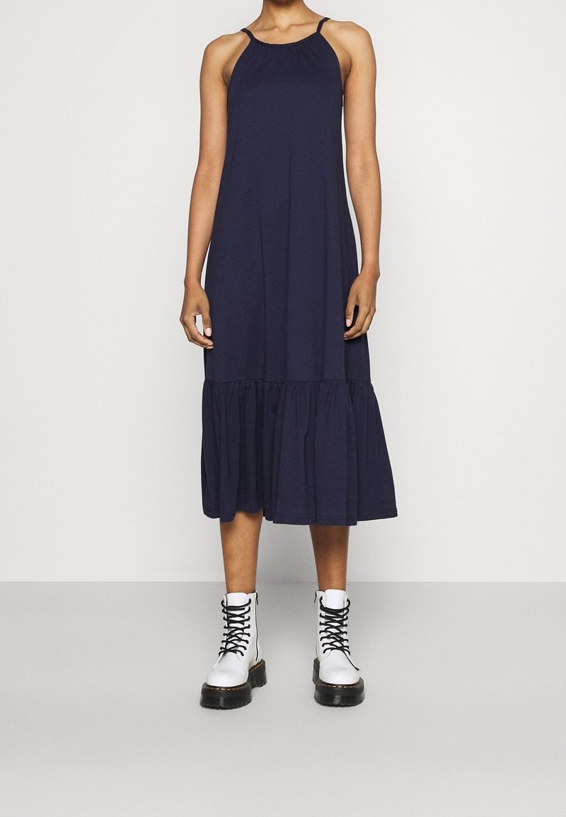 Mörkblå ärmlös klänning med en samlad byst och utflugen flerlagers nederkant, kombinerad med vita snörda kängor.