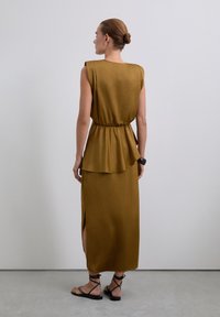 Femme avec un chignon portant une robe midi sans manches de couleur moutarde avec une fente sur le côté, des sandales noires et un bracelet noir, se tenant contre un mur blanc.