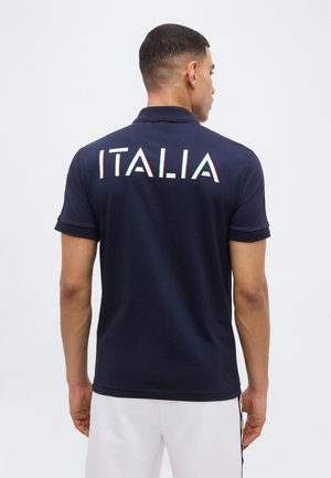 Vyras, atsisukęs nugara, dėvi tamsiai mėlyną polo marškinėlį su žodžiu „ITALIA“ baltomis, žaliai ir raudonai dažytomis raidėmis nugaroje, derinamas su baltais šortais.