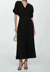 Donna che indossa un abito midi nero a pieghe con maniche a sbuffo corte, tiene in mano una borsa beige con borchie e indossa sandali neri con tacchi alti e cinturini.