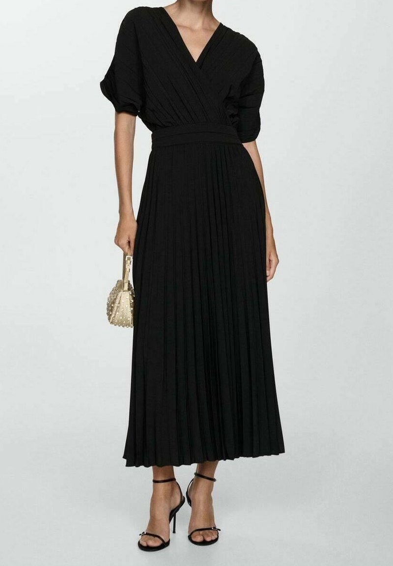 Donna che indossa un abito midi nero a pieghe con maniche a sbuffo corte, tiene in mano una borsa beige con borchie e indossa sandali neri con tacchi alti e cinturini.
