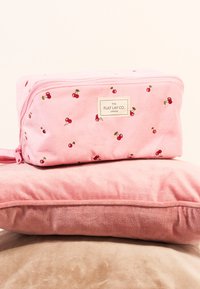 Roze stoffen cosmetica tas met kersenpatroon, ritsluiting en logo label. Geplaatst op zachte, pluche roze en beige kussens.