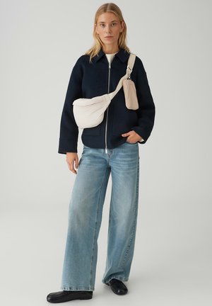 Femme portant une veste zippée bleu marine, un jean large bleu clair, des chaussures noires et un sac bandoulière crème, debout devant un fond uni.