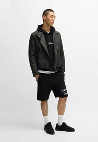 Giubbotto in pelle nera da motociclista, felpa nera con logo, pantaloni corti neri, calzini bianchi e scarpe da ginnastica nere. Texture liscia, design sportivo casual.