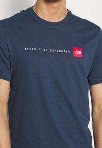 Camiseta azul marino de algodón, con el texto "NUNCA DEJES DE EXPLORAR" y un logo rojo en la parte izquierda del pecho. Mangas cortas.