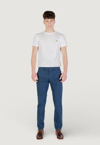 T-shirt bianco in cotone con maniche corte e logo verde, abbinato a pantaloni blu navy e scarpe da barca in pelle marrone.