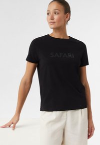 Czarny bawełniany T-shirt z napisem "SAFARI" w ciemnym, błyszczącym druku na przodzie. Krótkie rękawy i klasyczny dekolt typu crew neck.