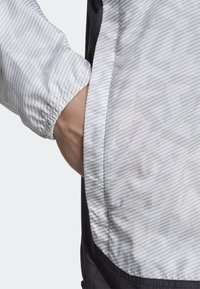 Lekka, biała kurtka w szare paski z elastycznymi mankietami i czarną dolną częścią; posiada boczną kieszeń na przechowywanie.