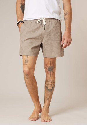 Homme portant un short beige texturé avec cordon de serrage, une chemise blanche, et des tatouages visibles sur les jambes et les bras, debout pieds nus sur un fond neutre.