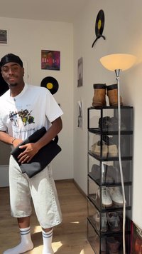 Jovem a usar um durag e uma t-shirt gráfica, segurando roupa preta dobrada perto da arrumação de sapatos e de uma lâmpada de pé numa divisão iluminada pelo sol, com decoração em vinil.