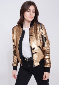 Goldene Pailletten Bomberjacke mit schwarzen Akzenten, ausgestattet mit einem Frontreißverschluss und Taschen, kombiniert mit einem grauen Shirt und schwarzen Jeans.