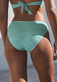 Lichtgroene bikini onderkant met een gestructureerd bloemenpatroon, voorzien van een low-rise snit en gladde randen. Verstelbare striksluiting aan de bovenkant.