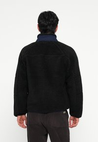 Schwarze Fleecejacke aus strukturiertem Material, mit hohem Kragen und blauer Innenseite. Verfügt über ein Markenlogo an der unteren Seite und hat einen lockeren Schnitt.
