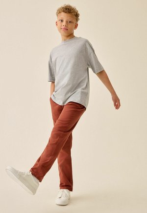 Dreng med krøllet hår iført en grå oversized t-shirt, rustfarvede bukser og hvide sneakers, stående med den ene fod let løftet.