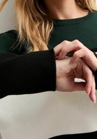Sweter robiony na drutach z zielonymi, czarnymi i białymi paskami; zbliżenie na teksturowany materiał i ręce z długimi paznokciami poprawiające mankiet rękawa.