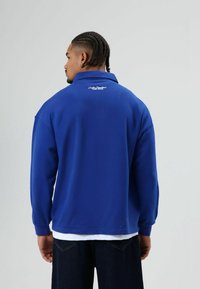 Blauwe trui met kraag, lange mouwen, zachte stof. Heeft geborduurde tekst op de achterkant, gedragen over een wit overhemd en donkere jeans.